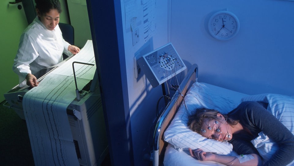 Frau liegt verkabelt im Schlaflabor, eine Ärztin im Nebenraum schaut auf Diagrammausdrucke.