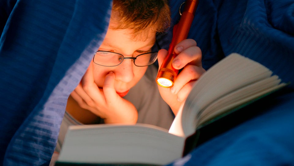 Kind liest im Bett ein Buch mit Taschenlampe.