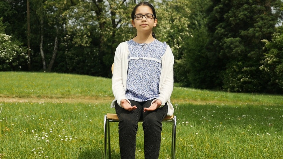 Mädchen sitzt mit geschlossenen Augen auf Stuhl und hat ihre Hände auf die Oberschenkel gelegt.