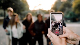 Im Vordergrund eine Hand, die ein filmendes Handy hält, im Hintergrund eine Gruppe von Jugendlichen auf der Straße.