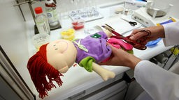 Hände halten Puppe über einem Labortisch und schneiden ihr mit Schere in die Hand.