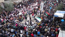 Syrische Demonstranten mit Flaggen und Plakaten stehen dicht gedrängt auf einem Platz.