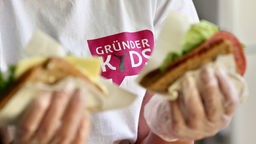Hände in Plastikhandschuhen halten geschmierte Brote, auf T'Shirt im Hintergrund steht 'Gründer Kids'.