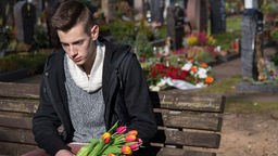 Teenager sitzt mit Blumen in der Hand auf einer Friedhofsbank.