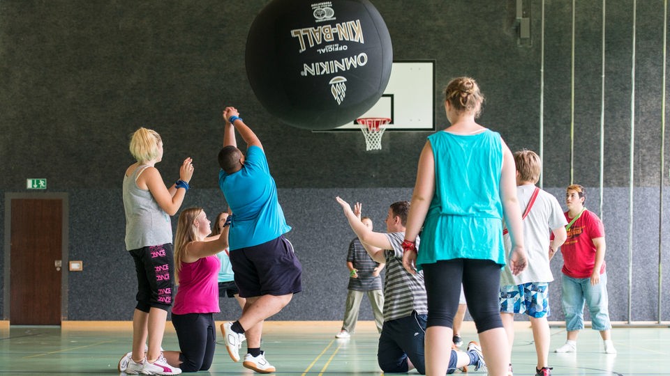 Übergewichtige Kinder spielen in Turnhalle mit riesigem Ball.