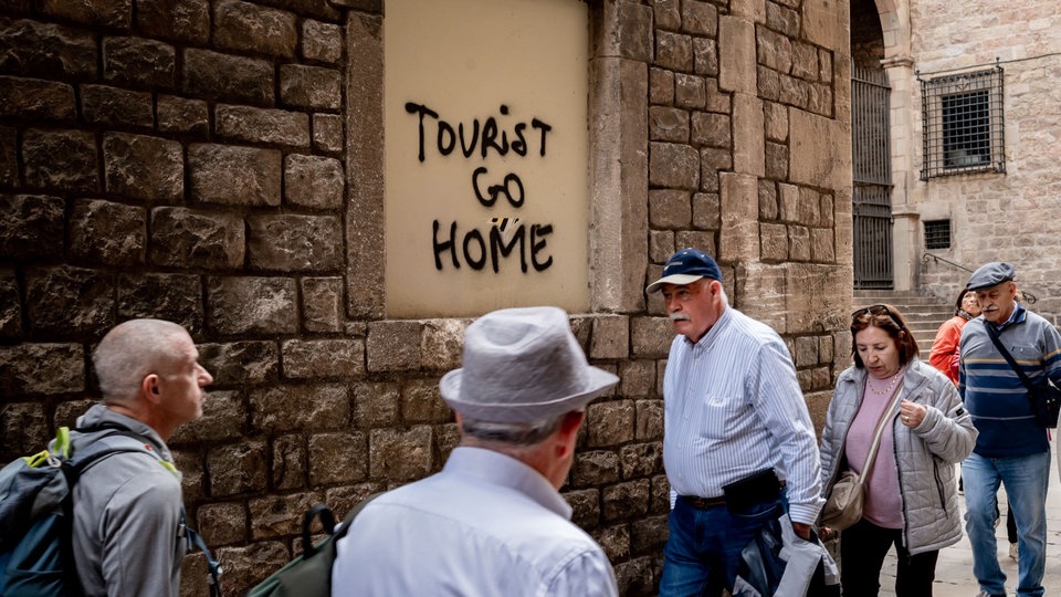 Menschen gehen an einer Wand entlang, auf der das Graffiti "Tourist go home" zu lesen ist.