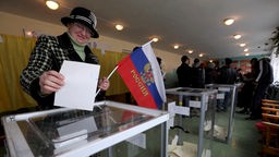 Wählerin wirft Wahlzettel in Urne und hält dabei eine russische Fahne in der Hand.
