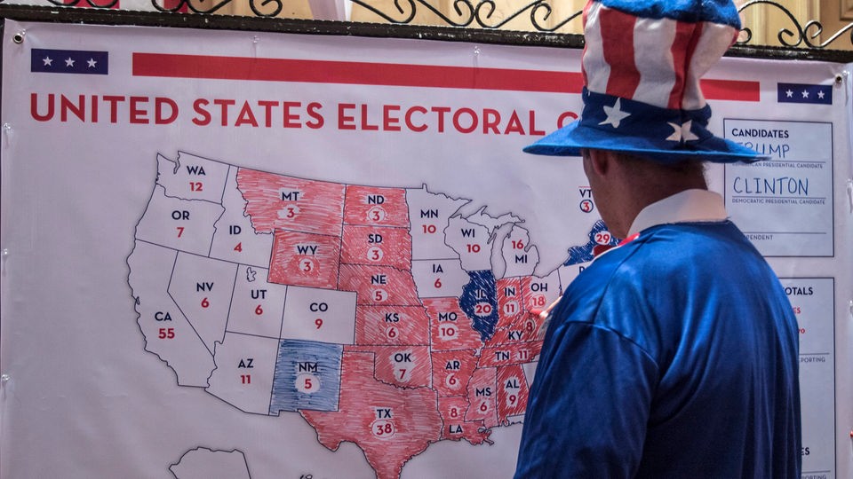 Mann mit Stars-and-Stripes-Hut malt auf US-Karte mit roten und blauen Bundesstaaten, um das Ergebnis der US-Präsidentschaftswahl zu dokumentieren.