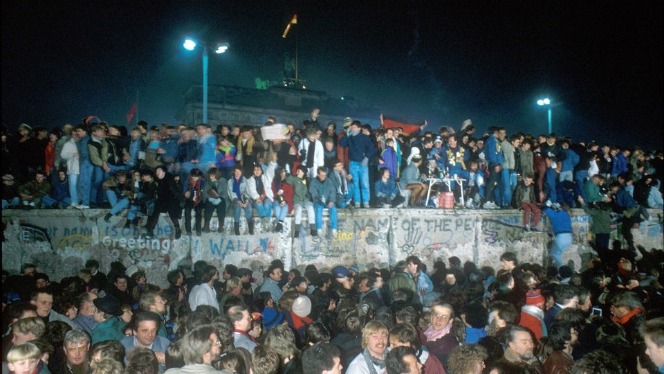 Jubelnde Menschen stehen auf der Mauer und feiern die deutsche Wiedervereinigung.