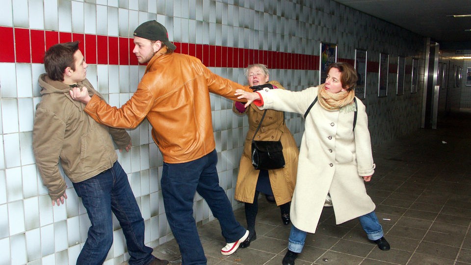 Zwei Frauen halten einen jungen Mann am Arm, der einen anderen schlagen will.