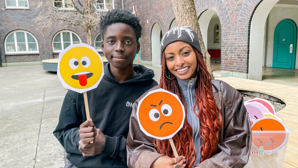 neuneinhalb-Reporterin Luam sitzt mit einem Schüler auf dem Pausenhof. Sie halten bunte Smileys mit verschiedenen Gesichtsausdrücken in den Händen.