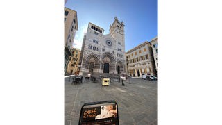 Auf einem Platz stehen vor einer Kirche Tische und Stühle, im Vordergrund Handy mit Anzeige 'Caffè al Tavolo'.