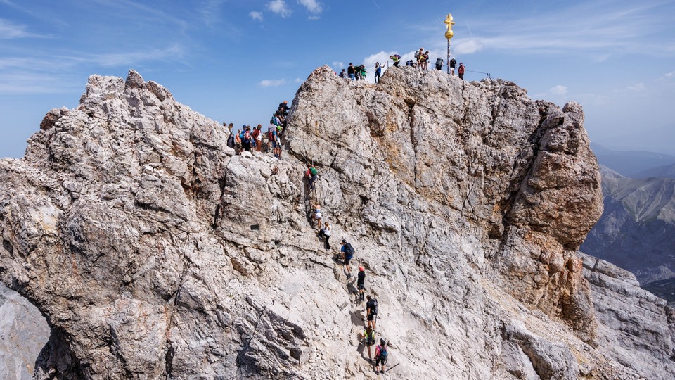 Zahlreiche Menschen klettern zum Gipfelkreuz der Zugspitze und stehen in Schlange an.