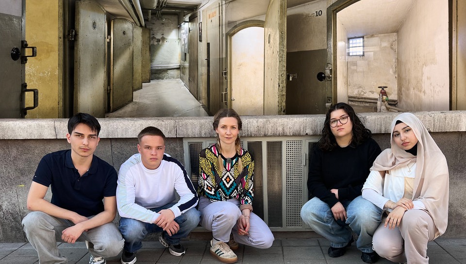 Reporterin Jana hockt zusammen mit vier Jugendlichen auf dem Boden vor einer Hauswand. Über ihnen ist ein Foto von Gefängniszellen projiziert, das aus dem NS-Dokumentationszentrum Kölns stammt.