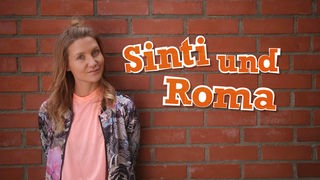 Jana steht vor einer Mauer. Neben ihr ist der Schriftzug „Sinti und Roma“ zu lesen.