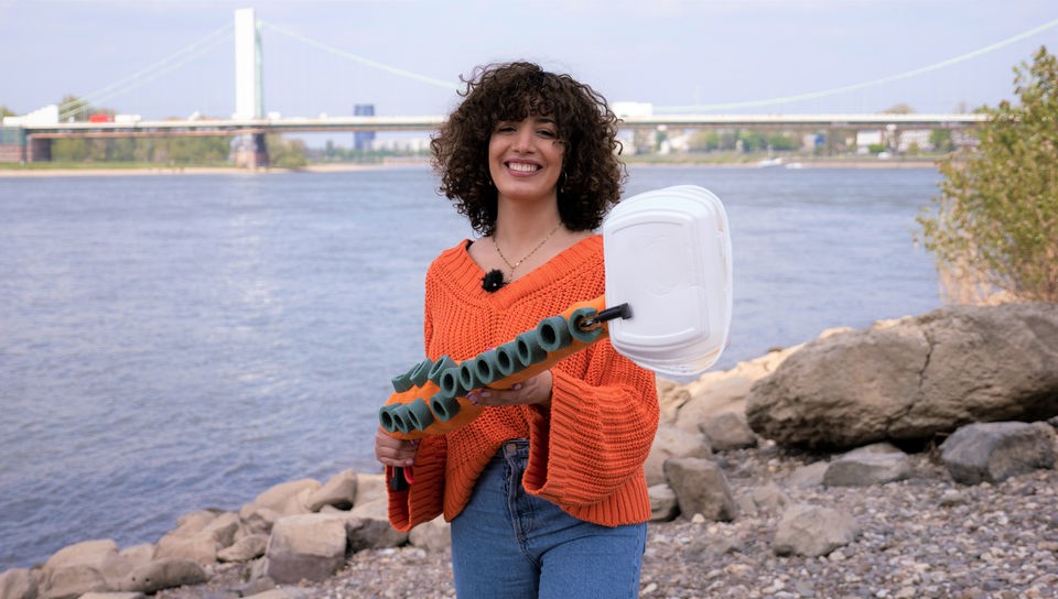 Reporterin Tessniem steht am Rheinufer mit einem Müllgreifer, der aussieht wie ein Tentakel.