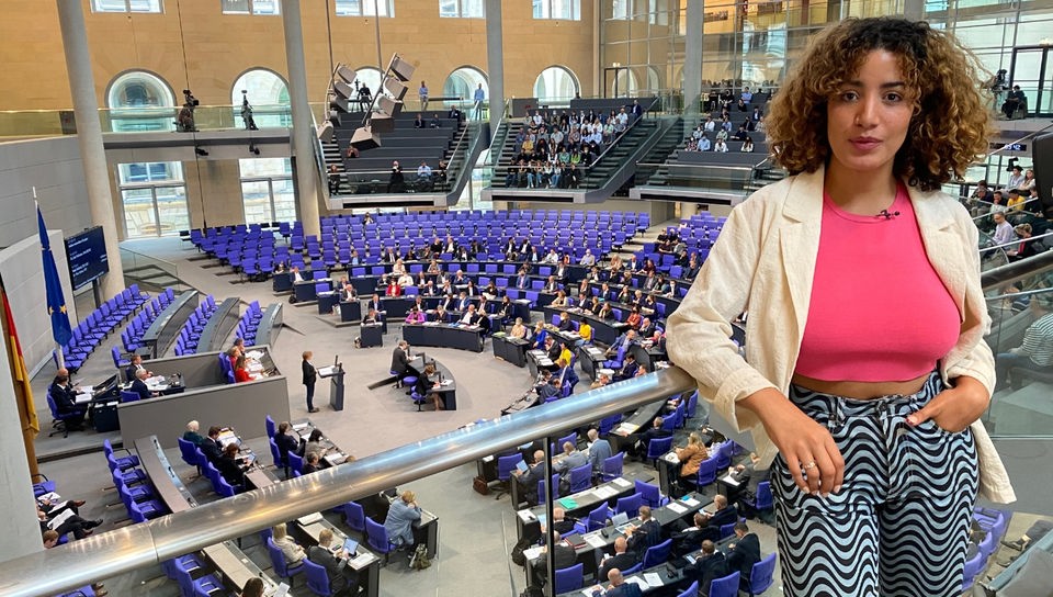 Reporterin Tessniem steht vor einer Glasabtrennung und schaut in die Kamera. Unter ihr ist der Plenarsaal mit Abgeordneten des Deutschen Bundestages.