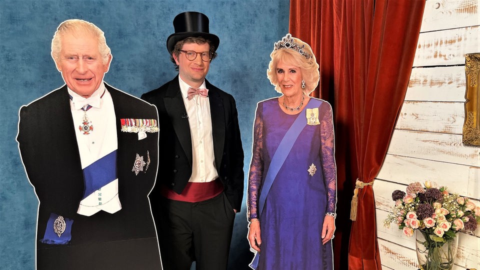 Reporter Robert hat sich schick gemacht und steht im Frack neben Pappaufstellern von Königin Camilla und König Charles III. im neuneinhalb-Studio.