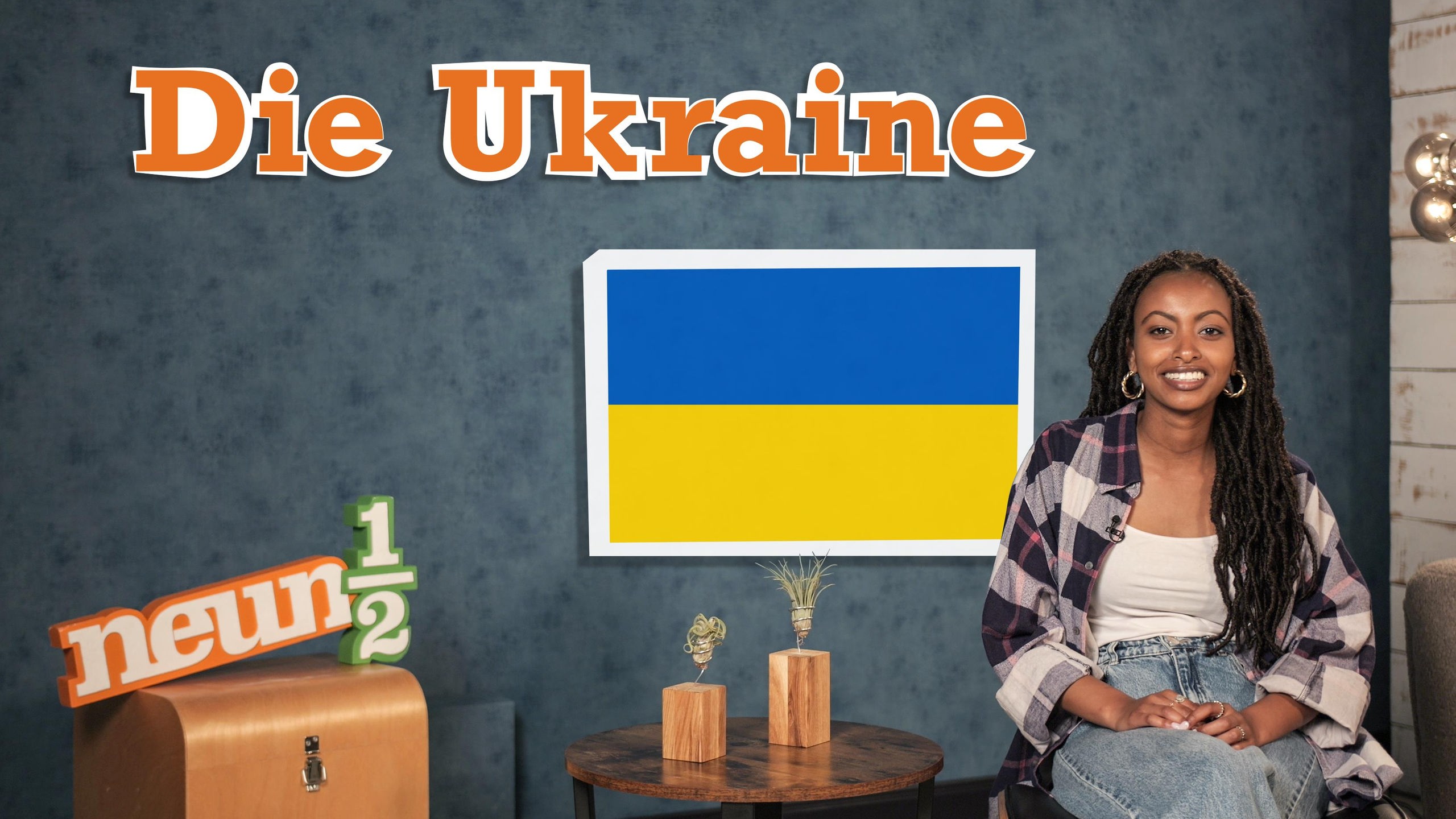 Luam sitzt auf einem Stuhl neben einem kleinen Tisch und dem neuneinhalb-Logo. Auf einer blauen Wand steht der Schriftzug „Die Ukraine“ und hinter Luam ist die ukrainische Flagge zu sehen.