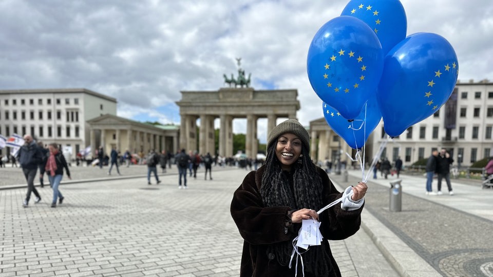 Reporterin Luam steht vor dem Brandenburger Tor und hält EU-Ballons in die Luft.