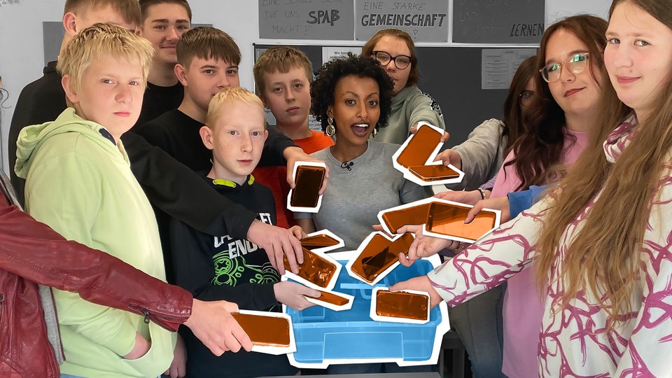 Reporterin Luam steht im Klassenzimmer und hält eine Kiste. Um sie herum stehen Schülerinnen und Schüler, die ihre Smartphones in die Kiste legen.