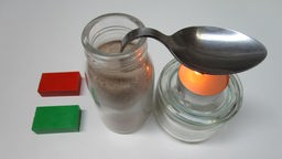 Gebogener Löffel steckt in sandgefülltem Glas, unter dem Löffel steht ein Teelicht.