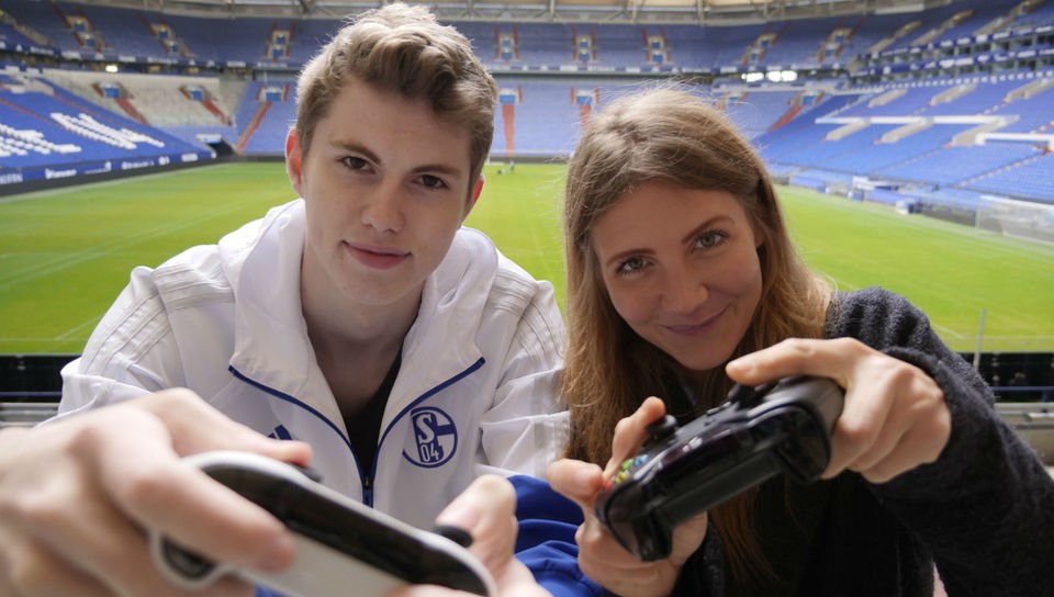 Jana und Tim Latka in einem leeren Fußballstadion, beide halten eine Fernbedienung für eine Spielekonsole.