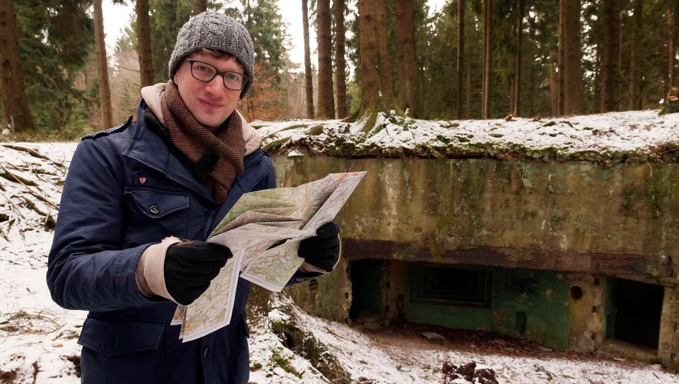 Robert steht mit Landkarte vor einem Bunker im Wald