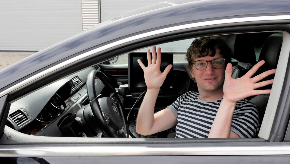 Robert sitzt in einem Auto, schaut aus dem Fenster und hebt die Hände vom Steuer.