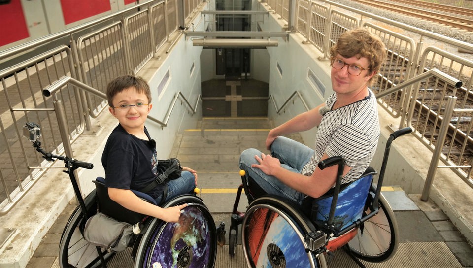 Robert und Timon stehen vor den Treppenstufen am Troisdorfer Bahnhof. Beide sitzen im Rollstuhl 