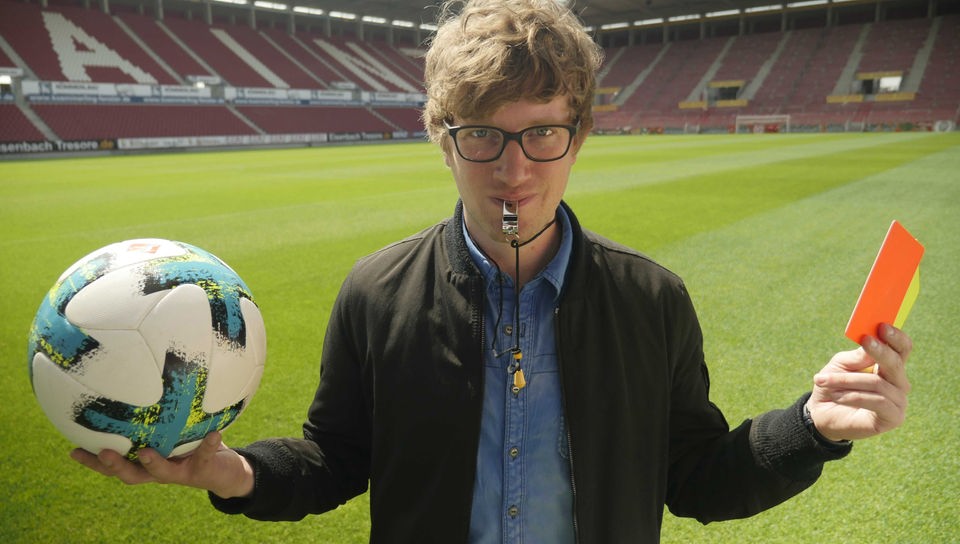 Robert steht mit Trillerpfeife im Mund auf einem Fußballfeld und hält einen Fußball und eine rote und gelbe Karte in den Händen.