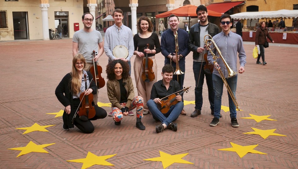 Siham mit den Musikern des Jugendorchesters der EU auf einem Platz, vor ihnen liegen gelbe Sterne auf dem Boden.