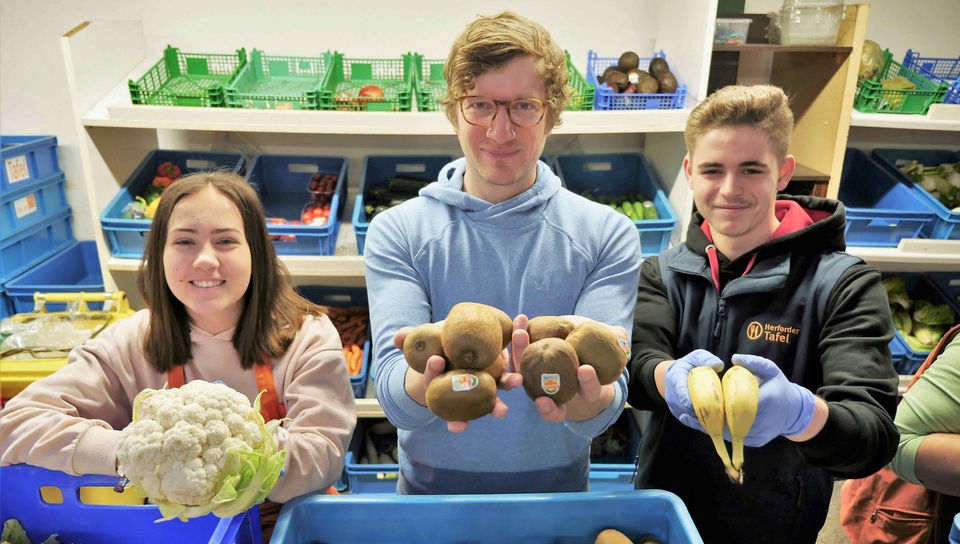 Robert zusammen mit Jacq und Lara, die bei der Herforder Tafel einen Bundesfreiwilligendienst leisten. Alle drei halten Obst und Gemüse in die Kamera.