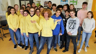 Gesa steht mit einer Schulklasse im Klassenzimmer. Eine Hälfte der Schüler trägt normale Klamotten, Gesa und die andere Hälfte tragen gelbe Sweatshirt-Jacken.