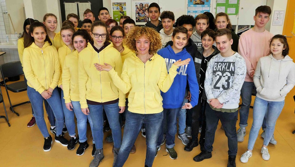 Gesa steht mit einer Schulklasse im Klassenzimmer. Eine Hälfte der Schüler trägt normale Klamotten, Gesa und die andere Hälfte tragen gelbe Sweatshirt-Jacken.