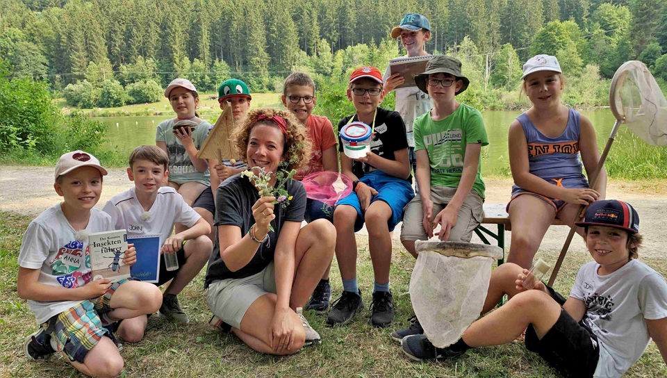 Gesa sitzt gemeinsam mit einer Gruppe junger Naturforscher*innen vor einem See. Die Kinder sind mit Käschern, Gefäßen und Büchern bewaffnet, Gesa hält Blumen in der Hand.