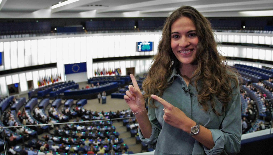 Mona steht auf der Empore des Straßburger Europaparlaments und zeigt auf den von Schüler*innen besetzten Plenarsaal. 
