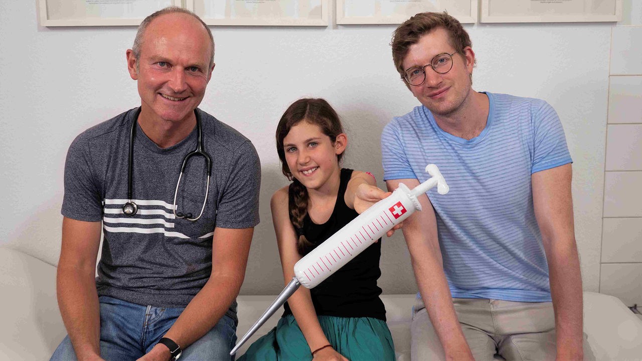 Robert sitzt neben dem Kinderarzt Dr. Höffer-Belitz und der 10-jährigen Lille. Lille hält eine große, aufblasbare Spritze in der Hand.