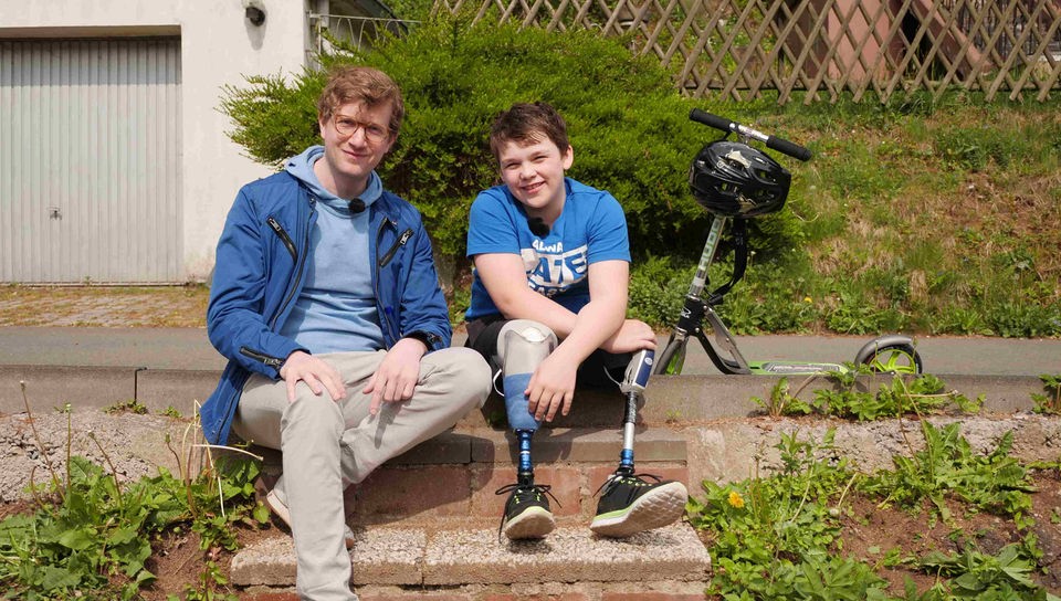 Robert sitzt neben dem 11-jährigen Elias draußen auf einer Steintreppe. Elias trägt eine kurze Hose, unter der seine zwei Beinprothesen zu sehen sind.