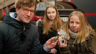 Robert steht gemeinsam mit den Schülerinnen Lara und Juliane vor einem Auto. In den Händen halten sie ein Stickoxid-Messgerät.