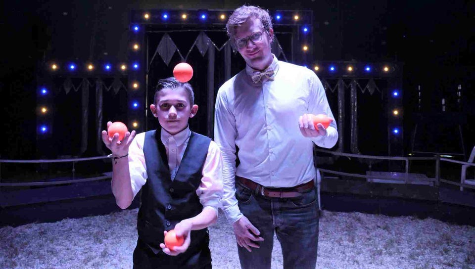 Robert und Miguel stehen in der Manege des Zirkus Amany. Miguel jongliert mit drei orangefarbenen Bällen.