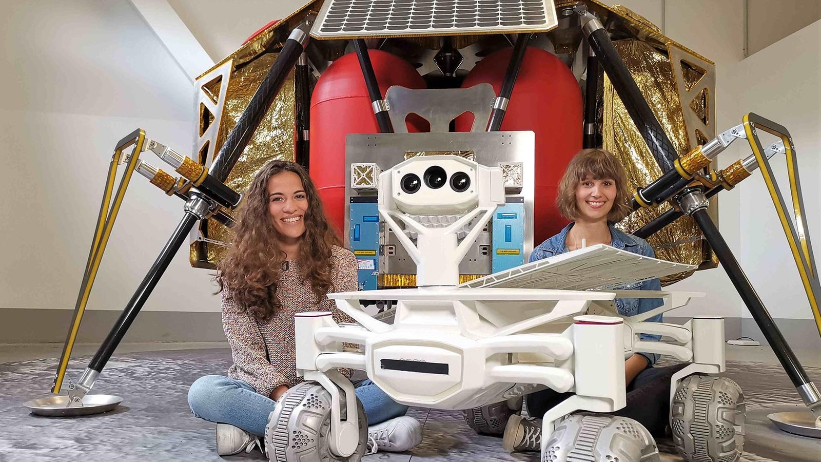 Mona und Wissenschaftlerin Nadine sitzen vor einer Raumsonde, neben ihnen steht ein Rover, der eines Tages auf dem Mond umherfahren soll. 