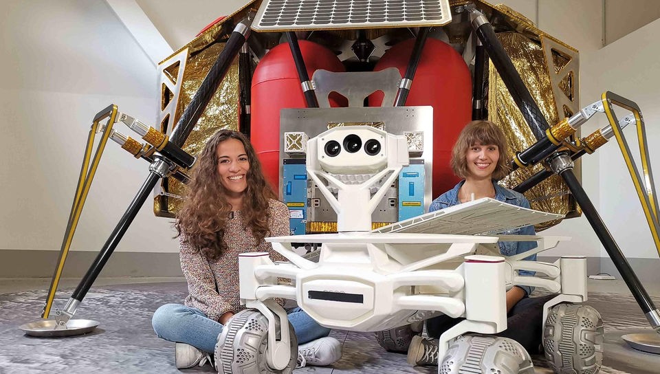 Mona und Wissenschaftlerin Nadine sitzen vor einer Raumsonde, neben ihnen steht ein Rover, der eines Tages auf dem Mond umherfahren soll. 