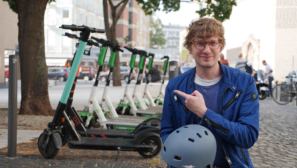 Robert steht in einer Fußgängerzone und trägt einen Fahrradhelm in der Hand. Mit der anderen zeigt er auf eine Reihe Elektro-Roller, die hinter ihm stehen.
