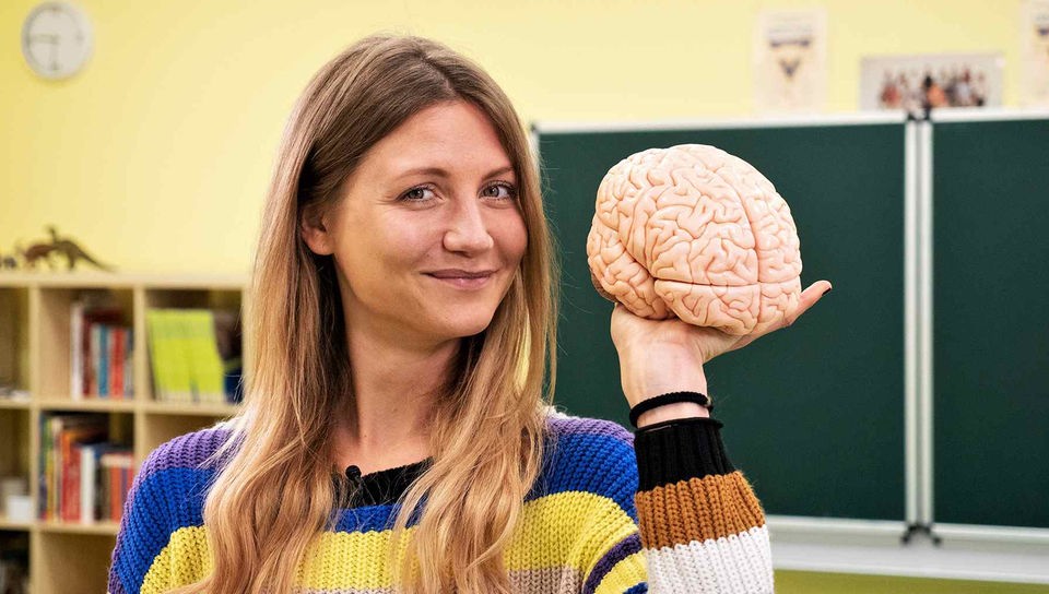 Jana steht in einem Klassenzimmer, sie hält das Modell eines Gehirns in der Hand.