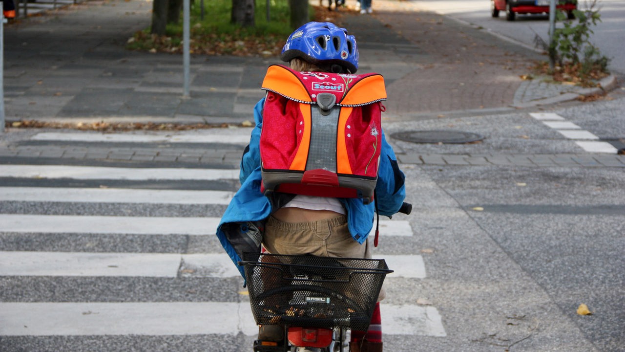 Kind mit Schulranzen und Helm steht mit dem Fahrrad an einem Zebrastreifen.