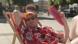 Robert schwitzt im sommerlichen Outfit in der Fußgängerzone der Stadt Bochum. Er sitzt in einem Sonnenstuhl, trägt ein Hawaiihemd und fächelt mit einem Handfächer.