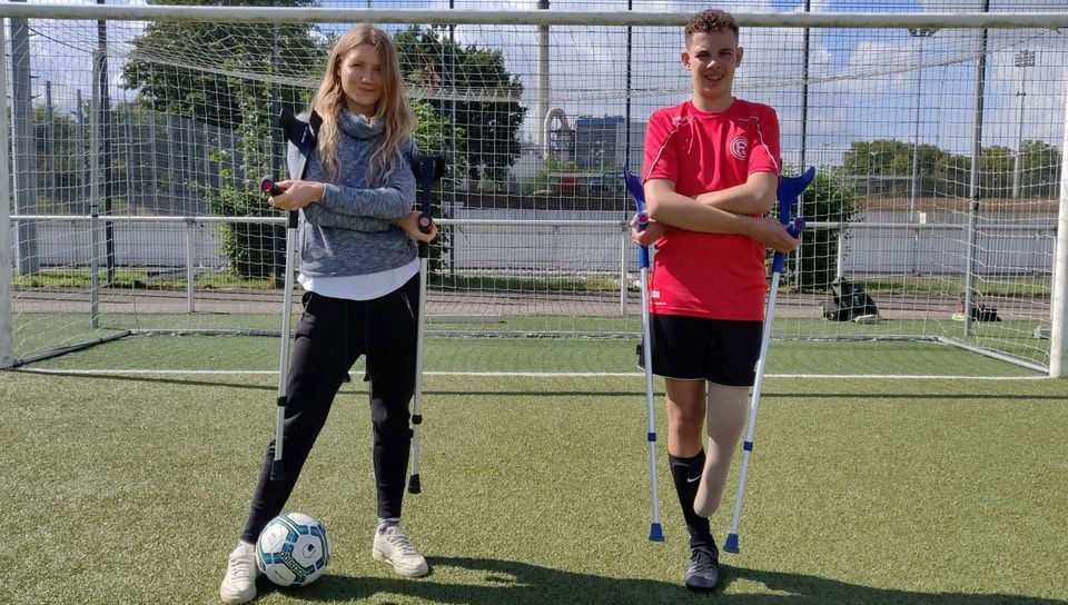 Reporterin Jana und der 14-Jährige Jamie stehen mit Krücken in den Händen vor einem Fußballtor. Jamie ist Amputierten-Fußballer, ihm fehlt ein Bein.