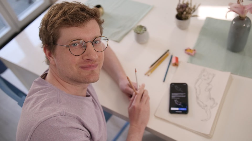 Reporter Robert sitzt an einem Tisch und hält mehrere Bleistifte in der Hand. Vor ihm liegt ein Blatt Papier mit einer Katzen-Zeichnung und ein Smartphone.