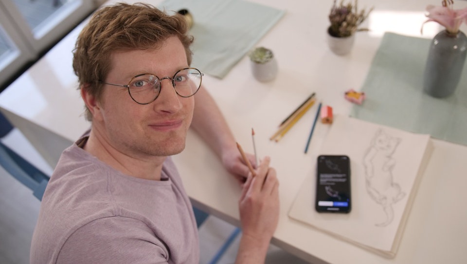 Reporter Robert sitzt an einem Tisch und hält mehrere Bleistifte in der Hand. Vor ihm liegt ein Blatt Papier mit einer Katzen-Zeichnung und ein Smartphone.